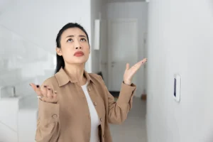 Homeowner standing near a wall thermostat and looking confused while troubleshooting an indoor comfort issue. Best for blogs about thermostat problems, HVAC not turning on, and cooling or heating issues.