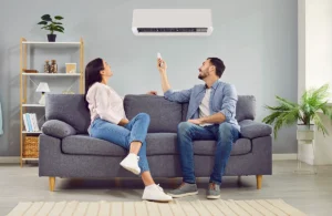 Couple sitting on a couch while using a remote to control a wall-mounted mini split air conditioner. Best for blogs about ductless mini splits, mini split operation, home cooling, and zoned comfort.
