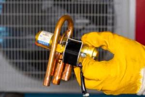 Close-up of a thermal expansion valve (TXV) assembly being held during HVAC repair or replacement. Best for blogs about reversing valve problems, heat pump repair, refrigeration components, HVAC part replacement, and why your heat pump is stuck in heating or cooling mode.