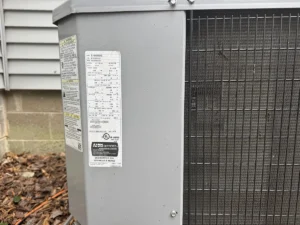 Close-up of the data label and condenser coil on an outdoor heat pump unit. Best for blogs about how to read HVAC model numbers, how to find tonnage on an AC unit, how old is my AC unit, HVAC serial number lookup, identifying your outdoor unit, and AC replacement planning.