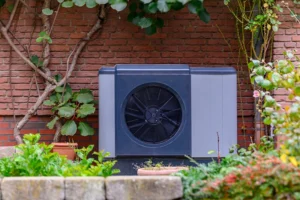 Outdoor heat pump unit installed in a landscaped yard near a brick wall.